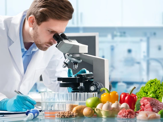 man in lab coat looks in microscope with various food items on table