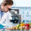 man in lab coat looks in microscope with various food items on table