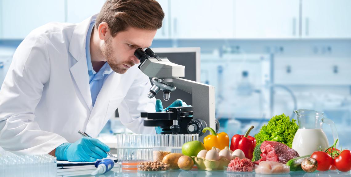 man in lab coat looks in microscope with various food items on table